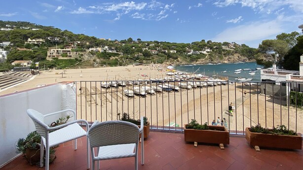 Un balcón con sillas de mimbre y macetas, con vista a una playa con barcos y un pueblo costero.