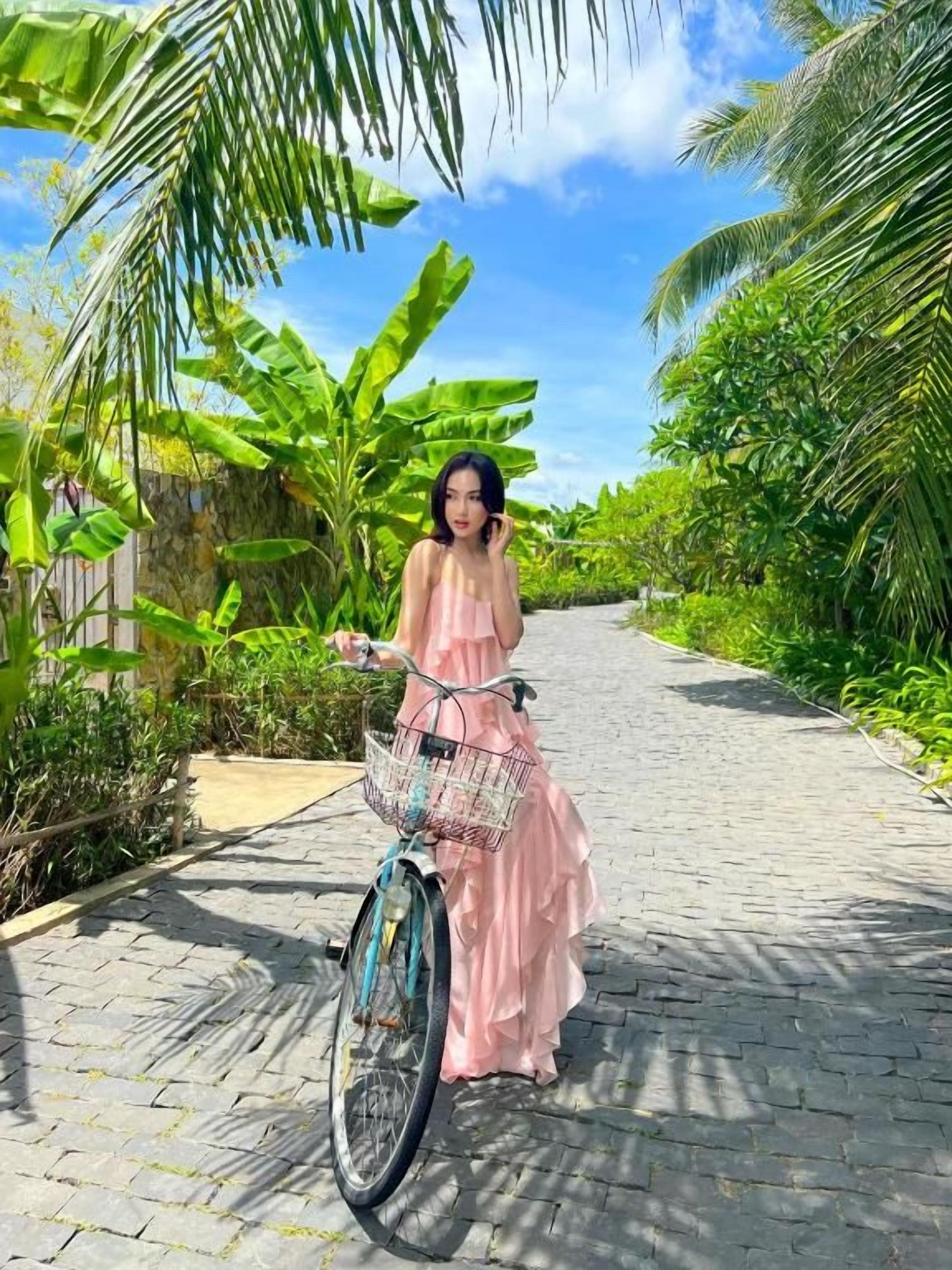 A person in a pink dress standing next to a bicycle with a basket, on a paved path surrounded by greenery.