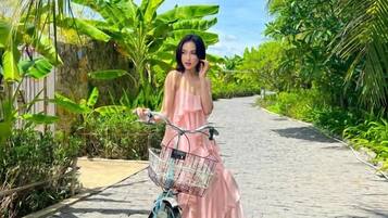 A person in a pink dress standing next to a bicycle with a basket, on a paved path surrounded by greenery.