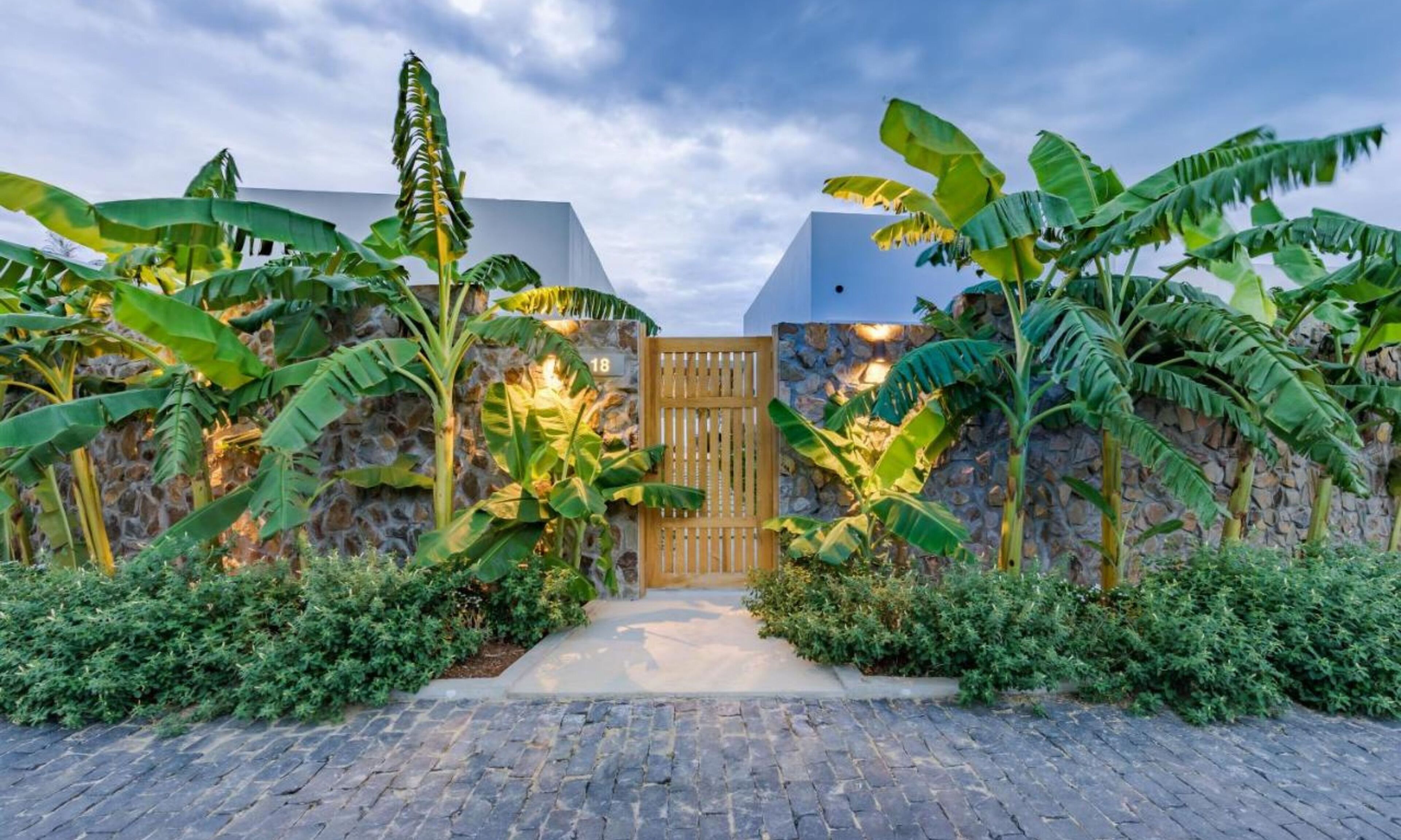Una entrada pavimentada con una puerta de madera, rodeada de exuberantes plantas de banano y un muro de piedra.