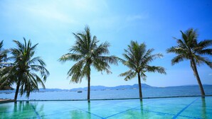 A clear blue pool with a tiled border, surrounded by palm trees and a view of the sea.