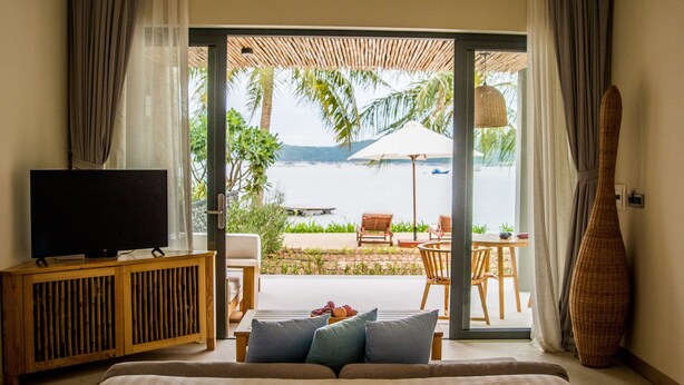 A room with a TV on a wooden cabinet, a sofa with cushions, a large window with a view of a beach and palm trees, and a wall with a textured design.