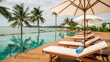 A wooden deck with lounge chairs and umbrellas overlooking a clear pool with palm trees and a distant view of mountains.