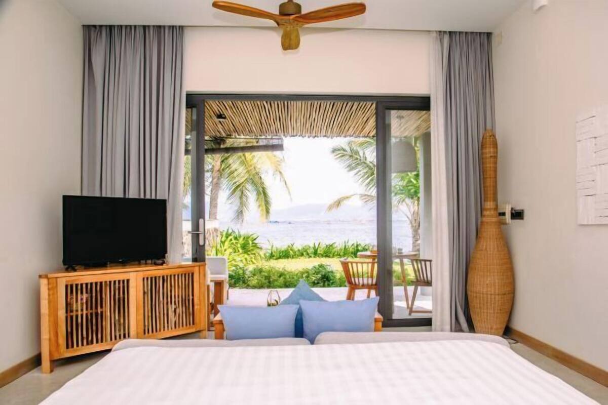 A bedroom with a bed, a TV on a wooden console, a ceiling fan, and a view of the ocean through a glass door.