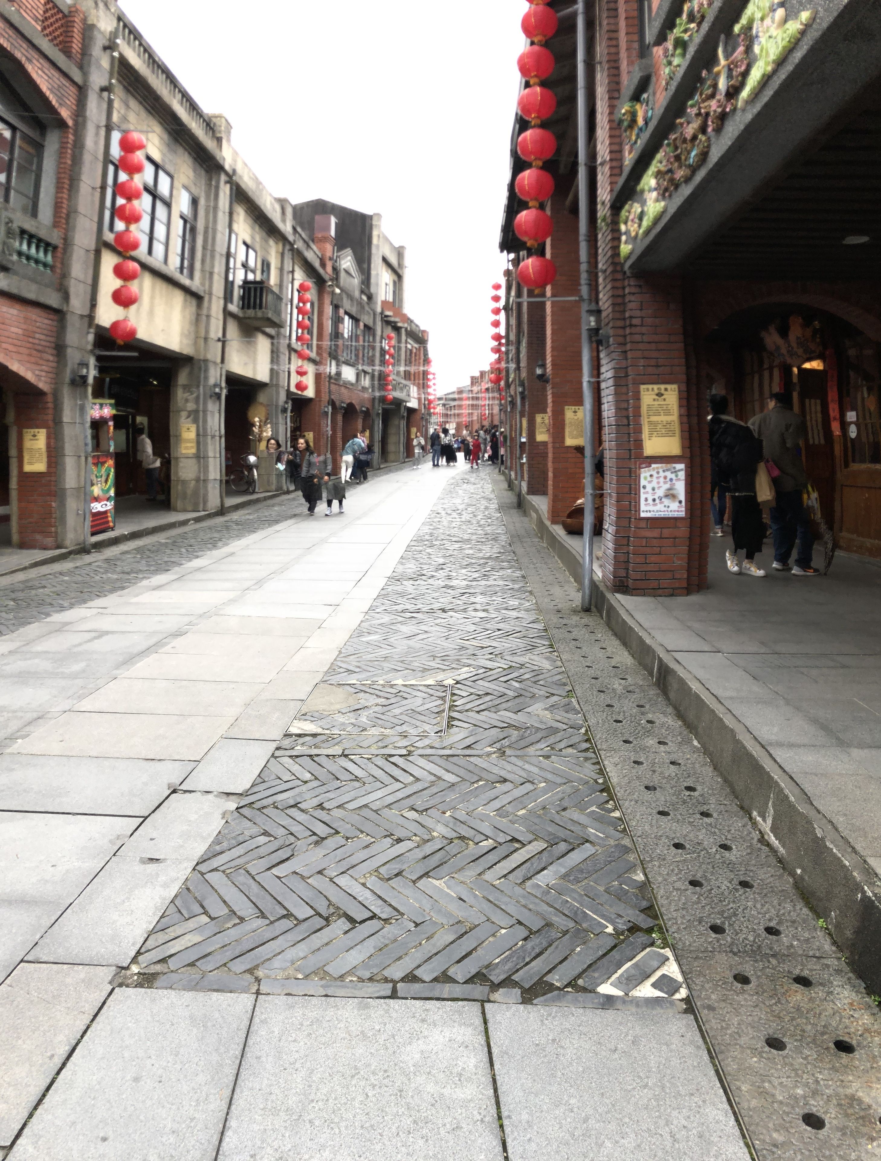 A cobblestone street with brick buildings, red lanterns, and a patterned pavement.