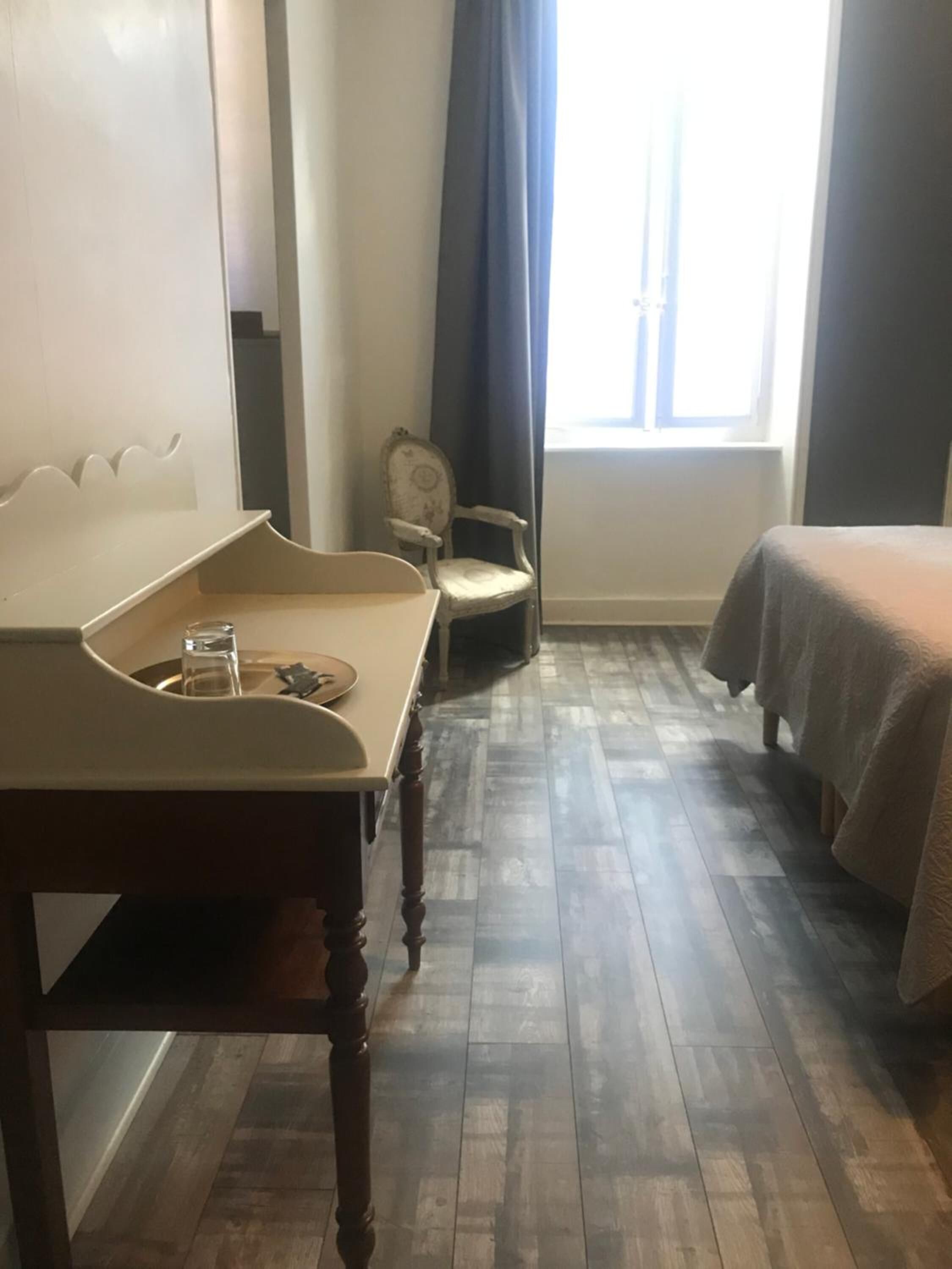Une pièce avec un plancher en bois, un lit recouvert d’une courtepointe blanche, une chaise et une petite table sur laquelle se trouvent un verre et une assiette.