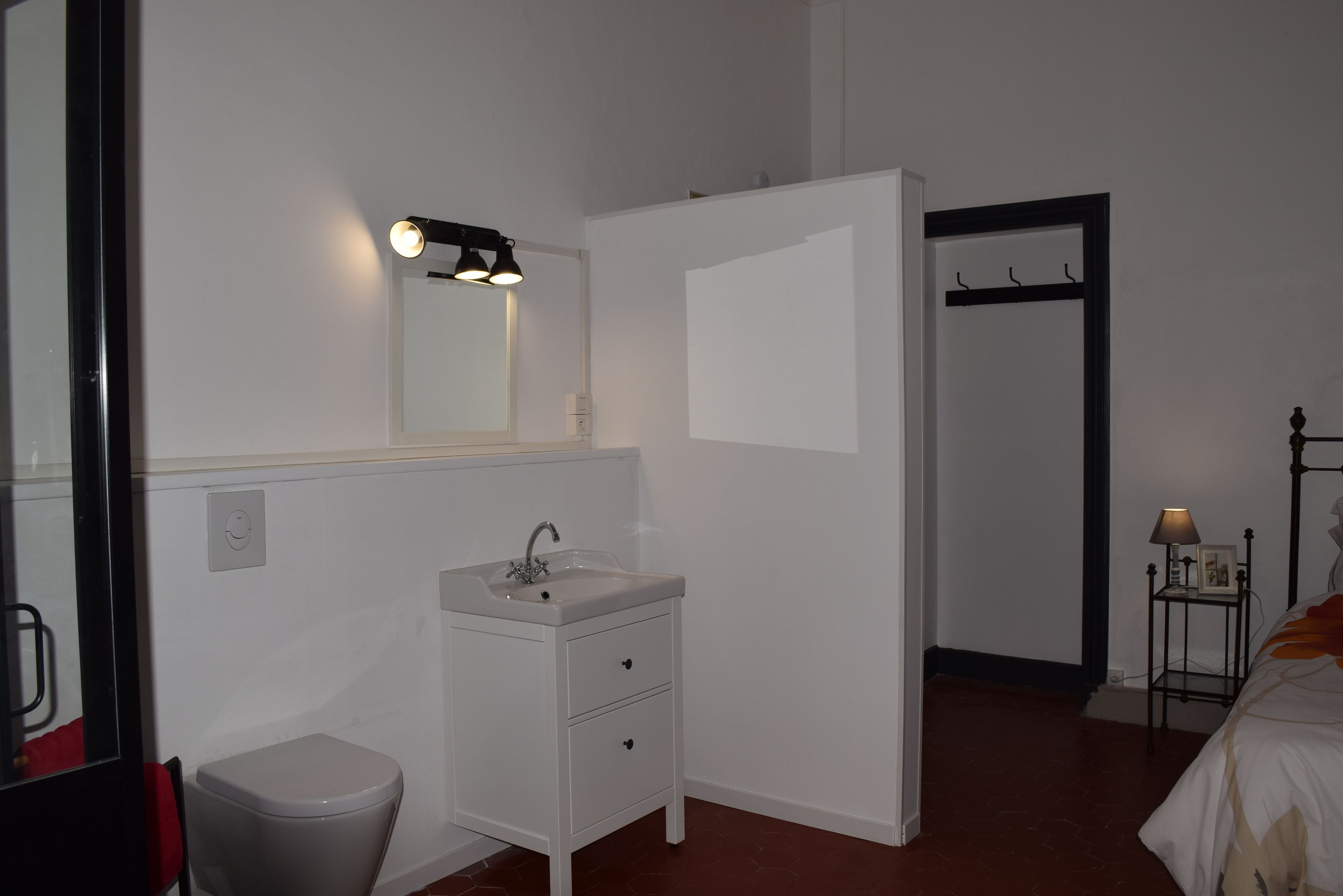 Une chambre d’hôtel avec un lavabo blanc, un miroir, une table de chevet avec une lampe, un lit et une porte donnant sur une autre pièce.