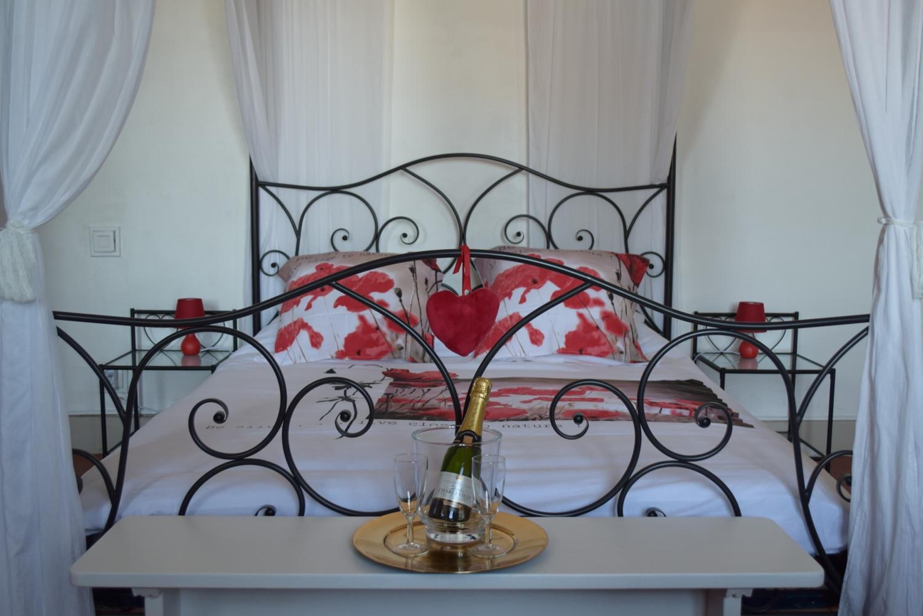A bed with a metal frame, white bedding, and a heart-shaped pillow. A bottle of champagne is placed on a tray on the bed.