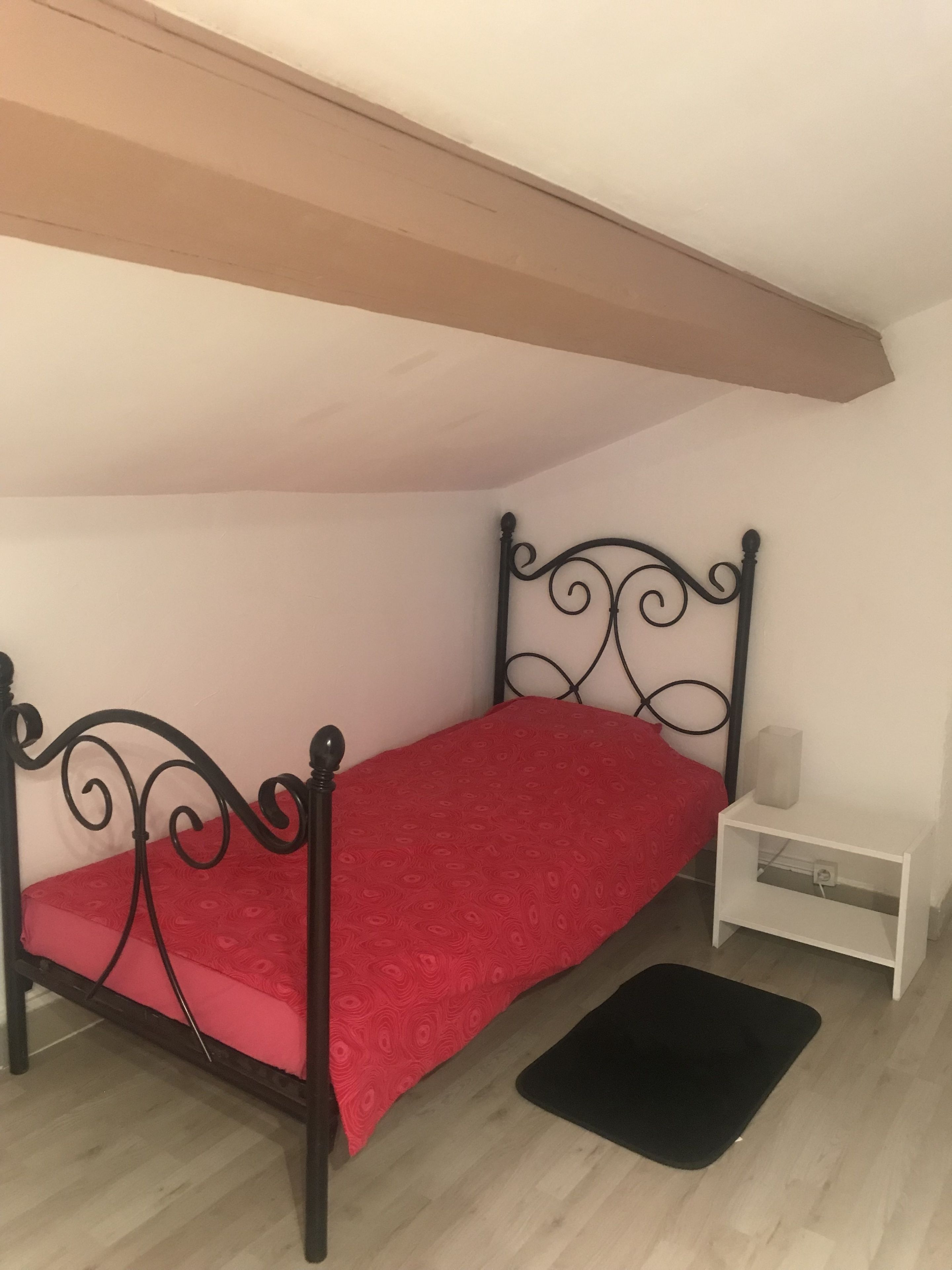 A small, single-bed room with a red bedspread, a black metal headboard, a white nightstand with a lamp, and a black mat on the floor.