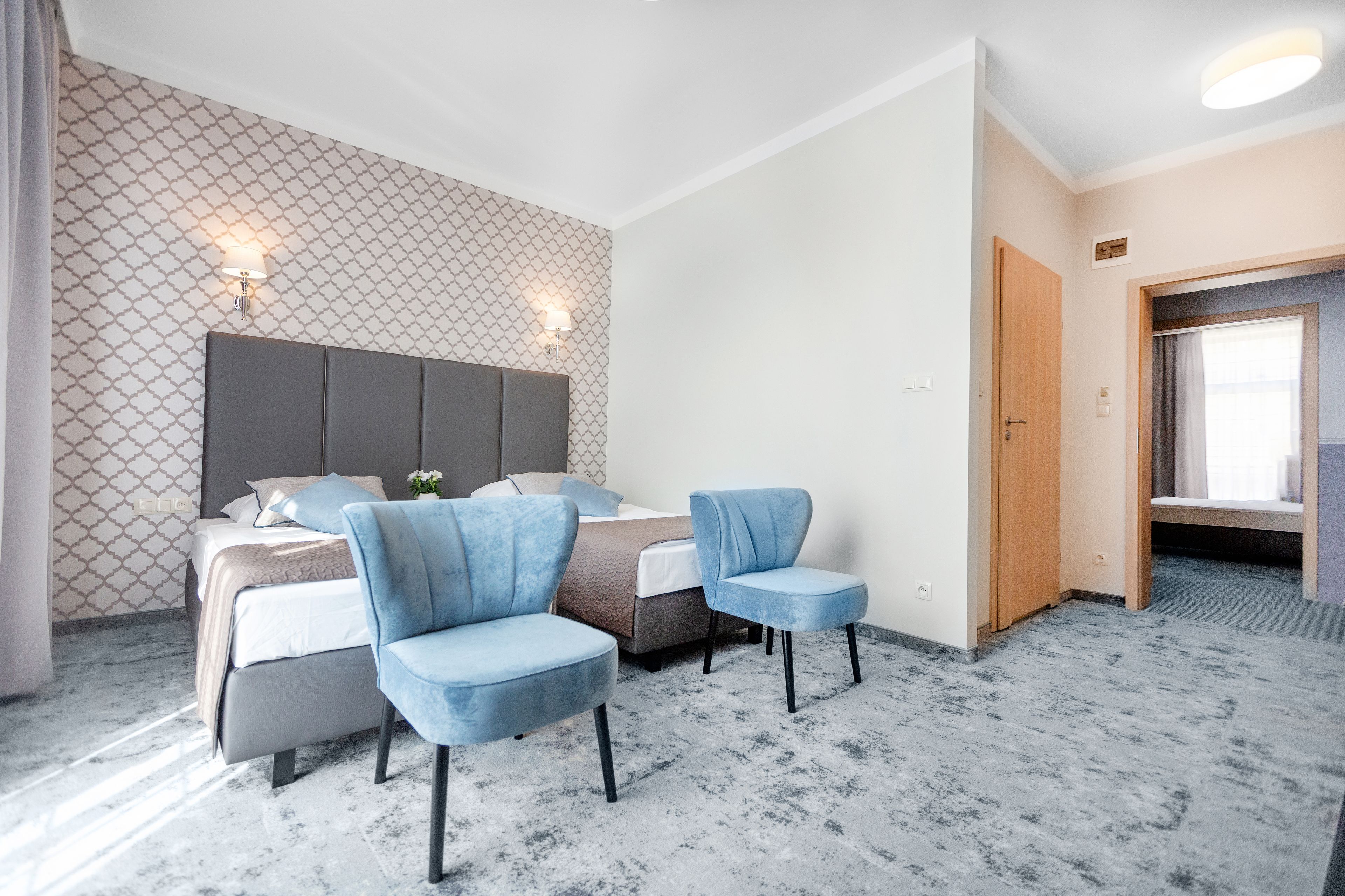 A hotel room with a bed, two blue chairs, a patterned wall, and a door leading to another room.