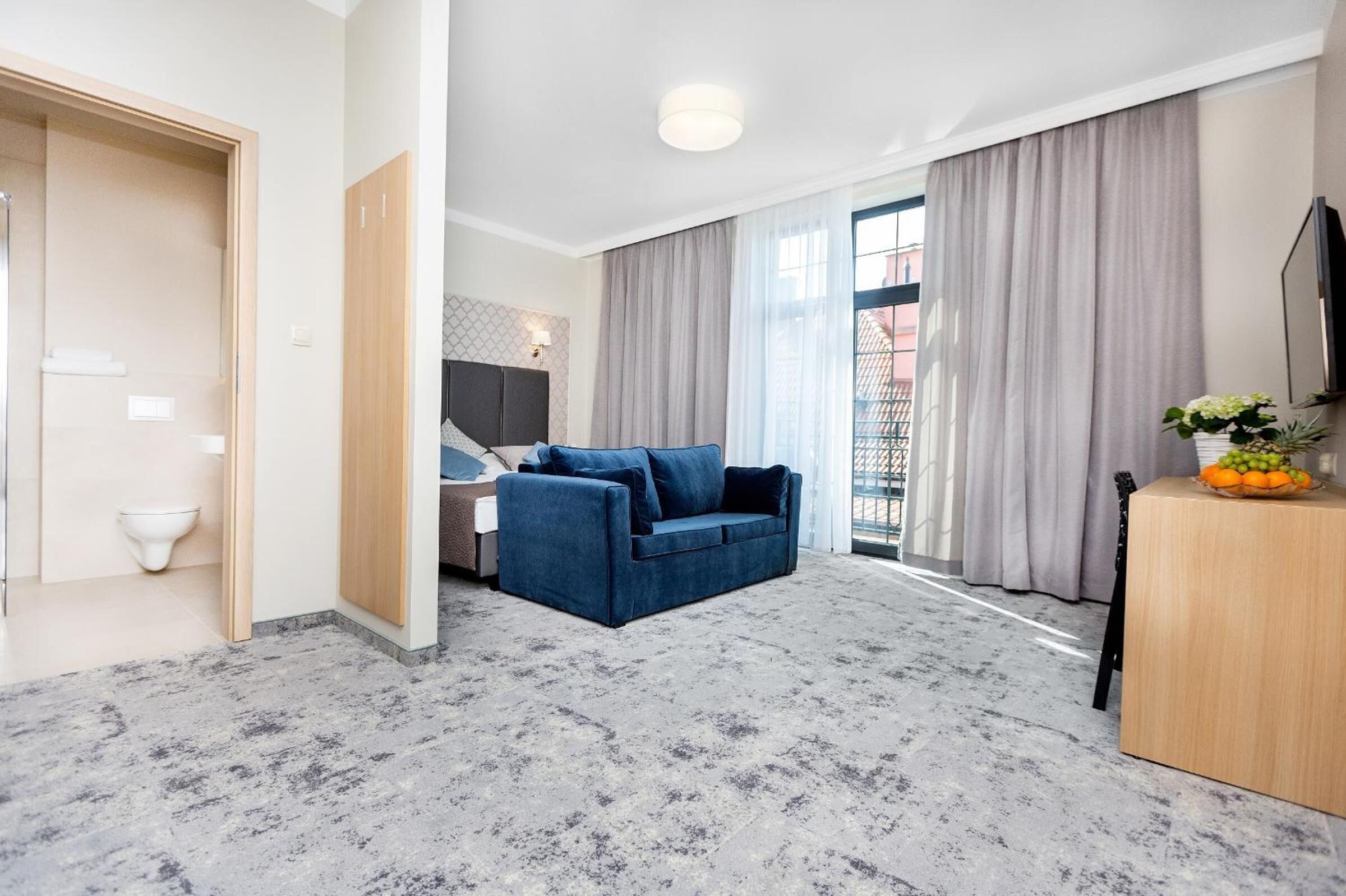 Quarto de hotel moderno com um sofá azul, uma escrivaninha de madeira e vista da cidade através de grandes janelas.