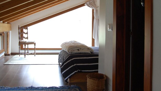 Un dormitorio con techo inclinado, una cama con ropa de cama a rayas, una silla y una ventana con cortinas.