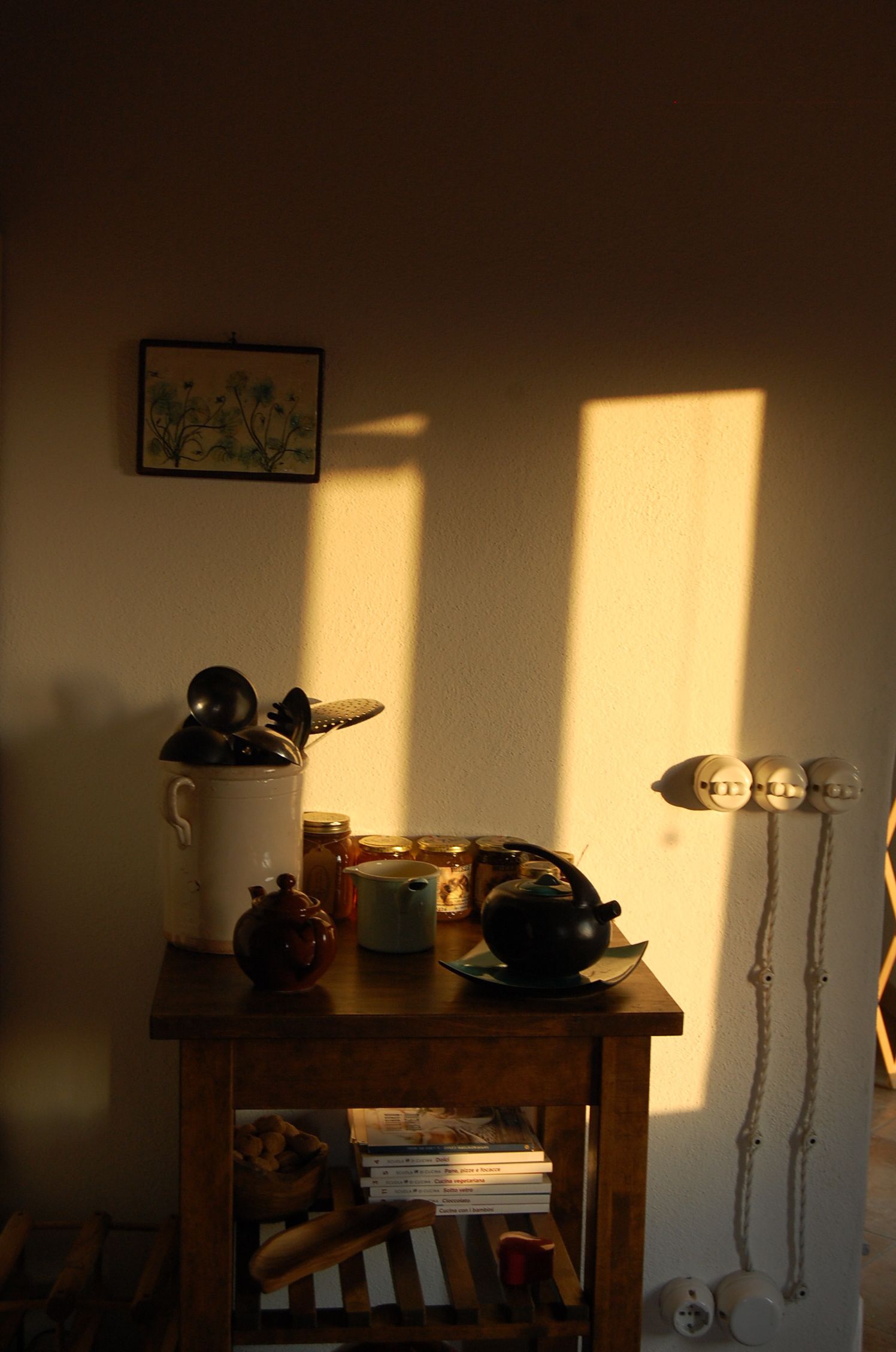 Una zona de cocina con una mesa de madera que sostiene utensilios de cocina, un hervidor y una taza. Hay frascos en la mesa y un cuadro en la pared.