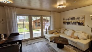 A living room with a sofa, a coffee table, a dining table, and a large window with a view of a house. - Prigge vacation apartment (Brodersby)