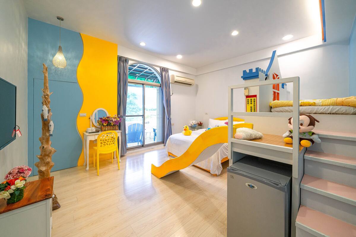 Habitación para niños con literas, escritorio, silla, refrigerador pequeño y ventana con cortinas.