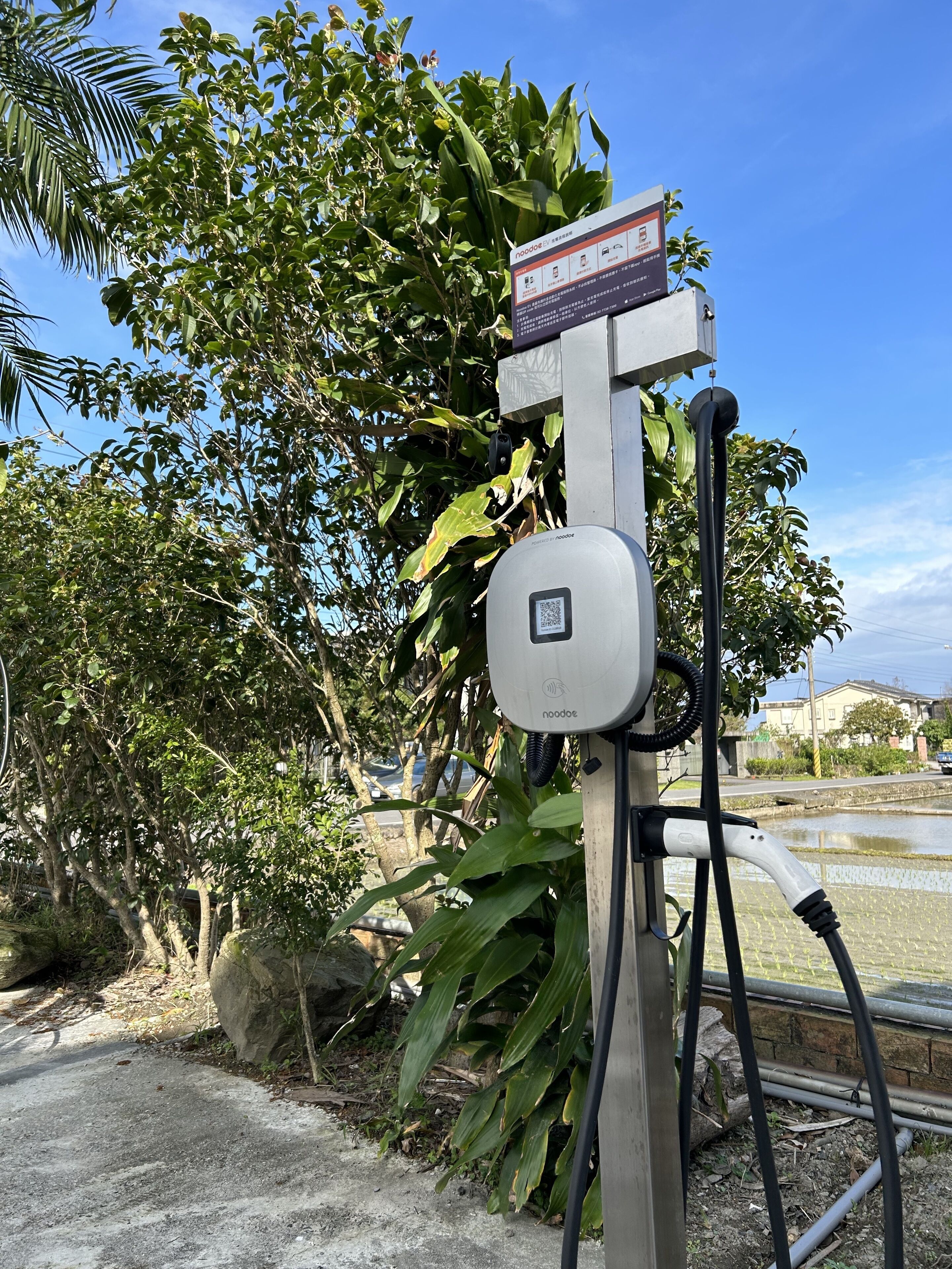 Una estación de carga para vehículos eléctricos con un código QR, rodeada de vegetación y un cielo despejado.