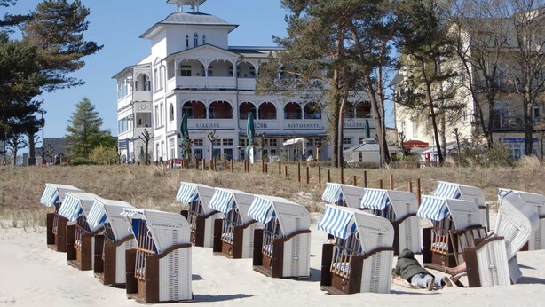A building with multiple floors, a prominent tower, and several signs indicating a restaurant, café, and pizzeria. Beach chairs are lined up in front of the building. - Holiday apartment 01 Villa Seeblick on the beach of Binz - Villa Seeblick Binz (Binz)