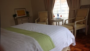A hotel room with a large bed, two armchairs, a small table with a drink, and a window with curtains. - Brooks (Nairobi)