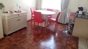 A dining area with a round table and red chairs, a white cabinet, and a kitchen area with a coffee machine.