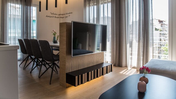 Ein modernes Wohnzimmer mit Flachbildfernseher, Essbereich mit Stühlen, Sofa und einer dekorativen Wandspruch.