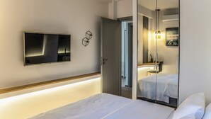 Ein modernes Hotelzimmer mit Bett, einem an der Wand montierten Fernseher, einem Nachttisch und einem Spiegel, der das Zimmer reflektiert.