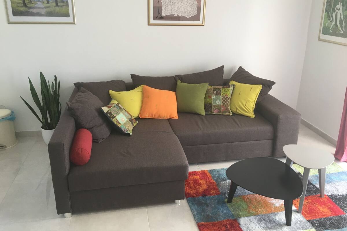 A living room with a brown sectional sofa, colorful pillows, a small round table, and a colorful rug. There are framed pictures on the wall and a potted plant on the floor.