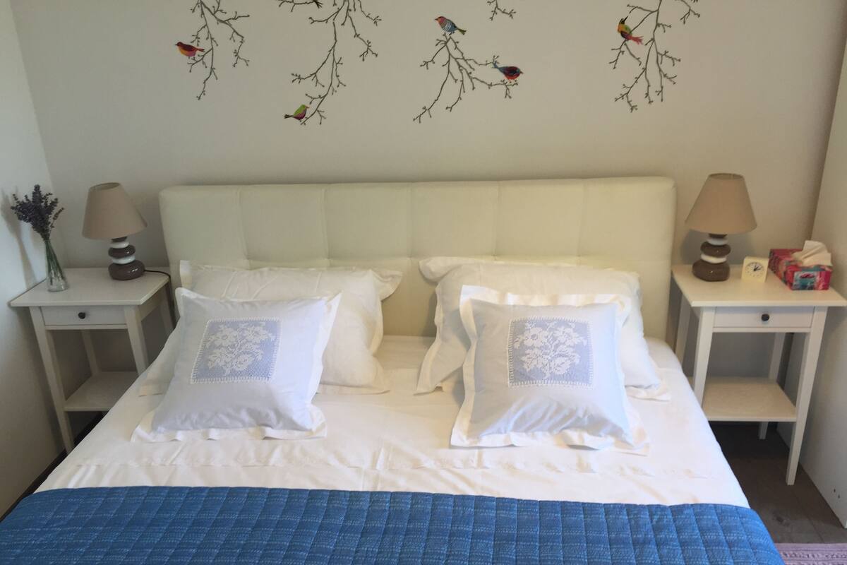 A neatly made bed with white bedding and blue quilt, flanked by two nightstands with lamps and a vase of flowers.