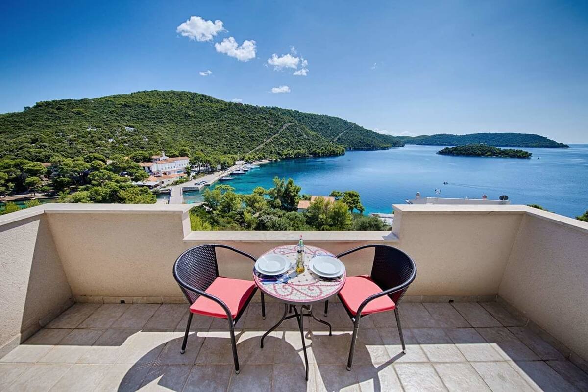 A balcony with two chairs and a table, overlooking a coastal area with a clear view of the sea and a green hillside.