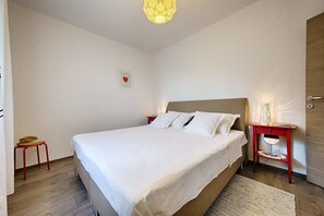 A neatly made bed in a room with a wooden floor, a small stool, a bedside table with a lamp, and a door.
