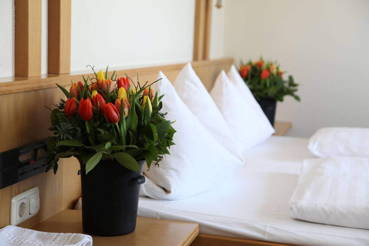 Habitación de hotel con cama, almohadas blancas, cabecera de madera y una planta con tulipanes rojos, amarillos y anaranjados.