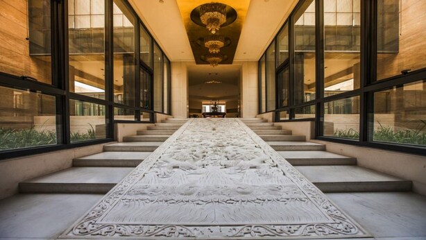 Un vaste hall d’entrée, avec un long tapis blanc aux motifs complexes qui mène à un escalier.