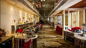 Un long couloir étroit d’hôtel, avec un tapis à motifs, des tables recouvertes de nappes rouges et un coin bar doté d’un grill.