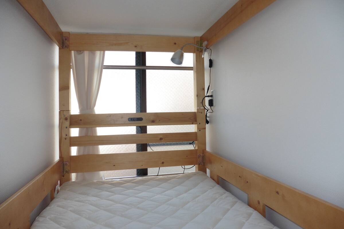Une petite chambre avec un lit simple et une structure de lit superposé en bois, une fenêtre avec des rideaux et une lampe fixée au mur.