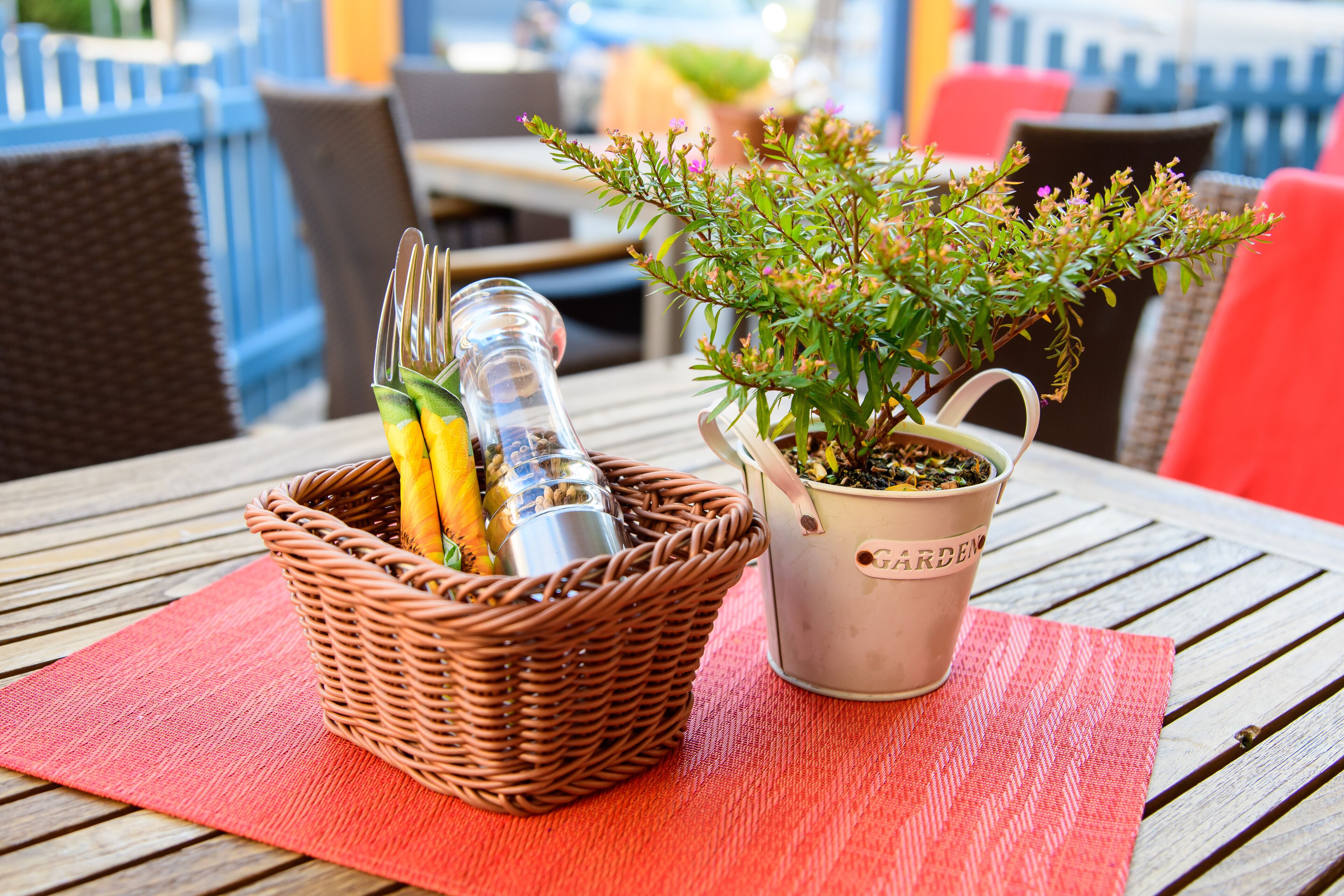 Una cesta de mimbre con utensilios y una planta en maceta sobre una mesa de madera con un mantel individual rojo.