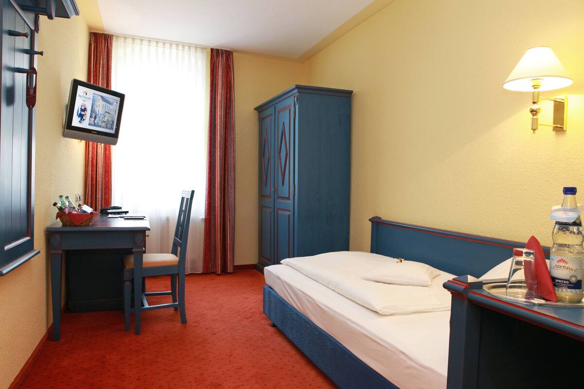 Une chambre d’hôtel comprenant un lit, un bureau, une chaise, une armoire, une table de chevet avec une bouteille et une lampe fixée au mur.