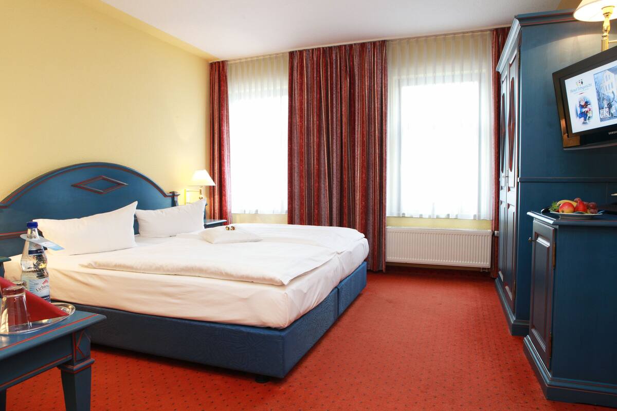 Une chambre d’hôtel avec un grand lit, un bureau avec une lampe, un téléviseur fixé au mur et une fenêtre avec des rideaux.