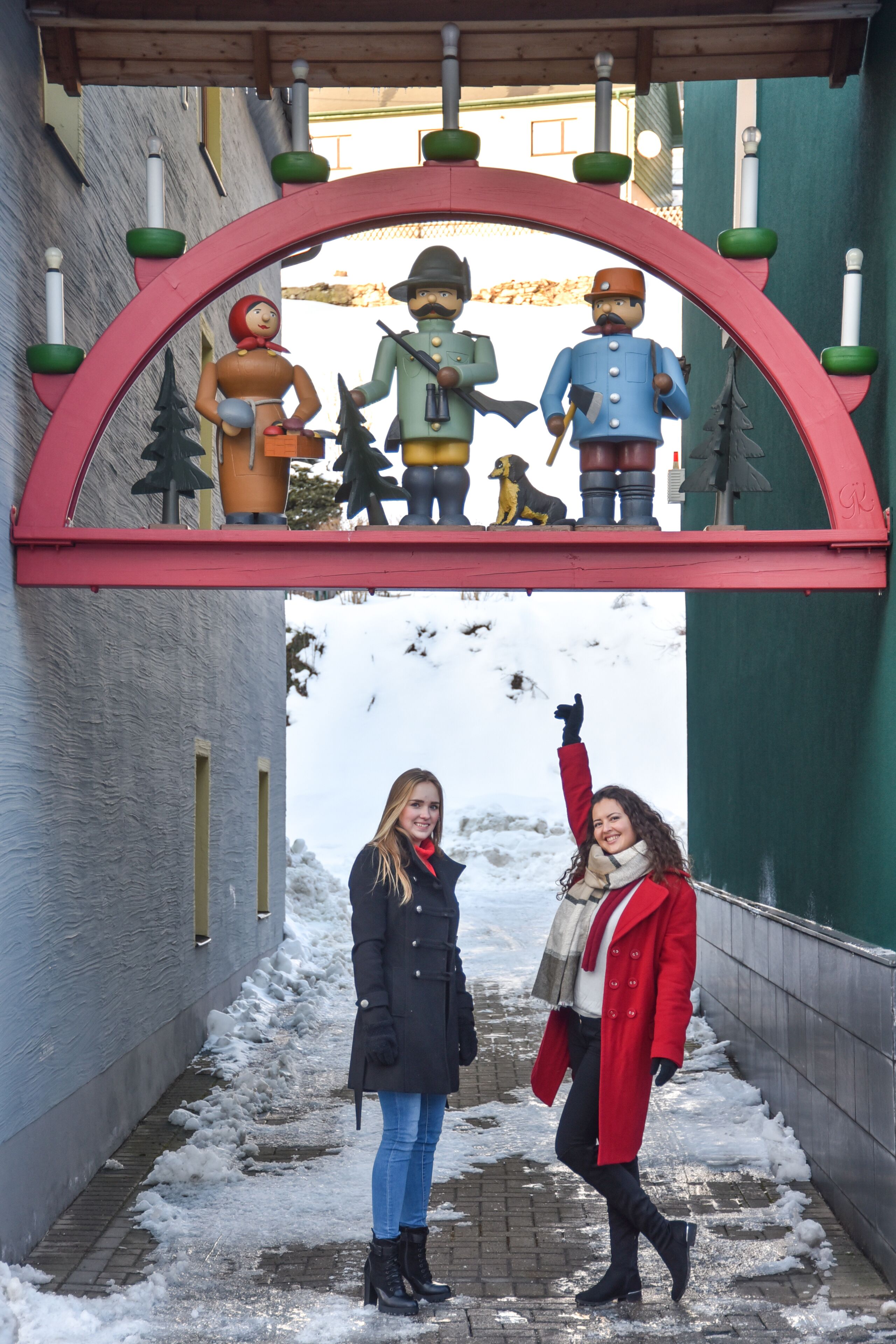 A narrow alley with snow on the ground, a red archway with figures, and two people walking.