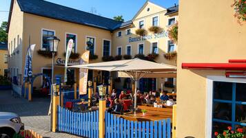 An outdoor seating area with tables and chairs, a blue fence, and a building with the sign "Buntes Haus Selbst".