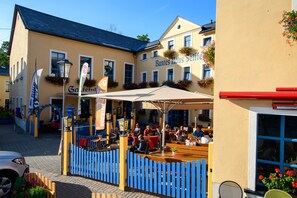 Ein Sitzbereich im Freien mit Tischen und Stühlen, ein blaues Geländer und ein Gebäude mit der Aufschrift 'Buntes Haus Selbst'.