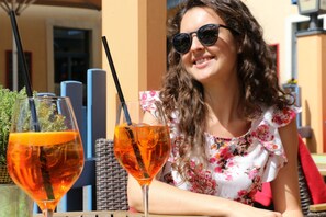 Eine Person sitzt an einem Tisch im Freien mit zwei Getränken, eines im Glas, das andere im Tumbler, mit einem Korbstuhl und einem roten Kissen im Hintergrund.