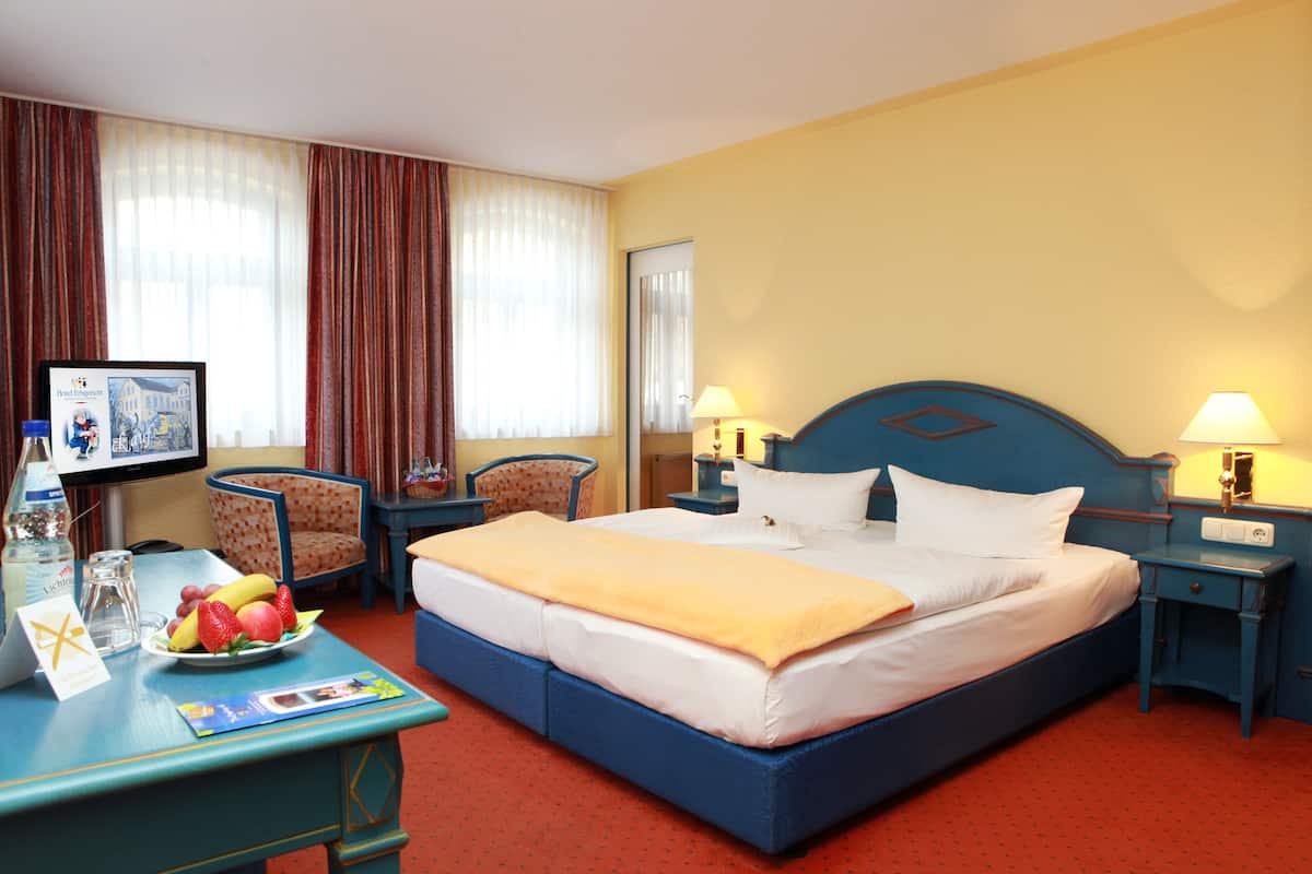 Une chambre d’hôtel avec un grand lit, deux tables de chevet avec des lampes, un bureau avec un ordinateur et un panier de fruits, une chaise et un téléviseur.