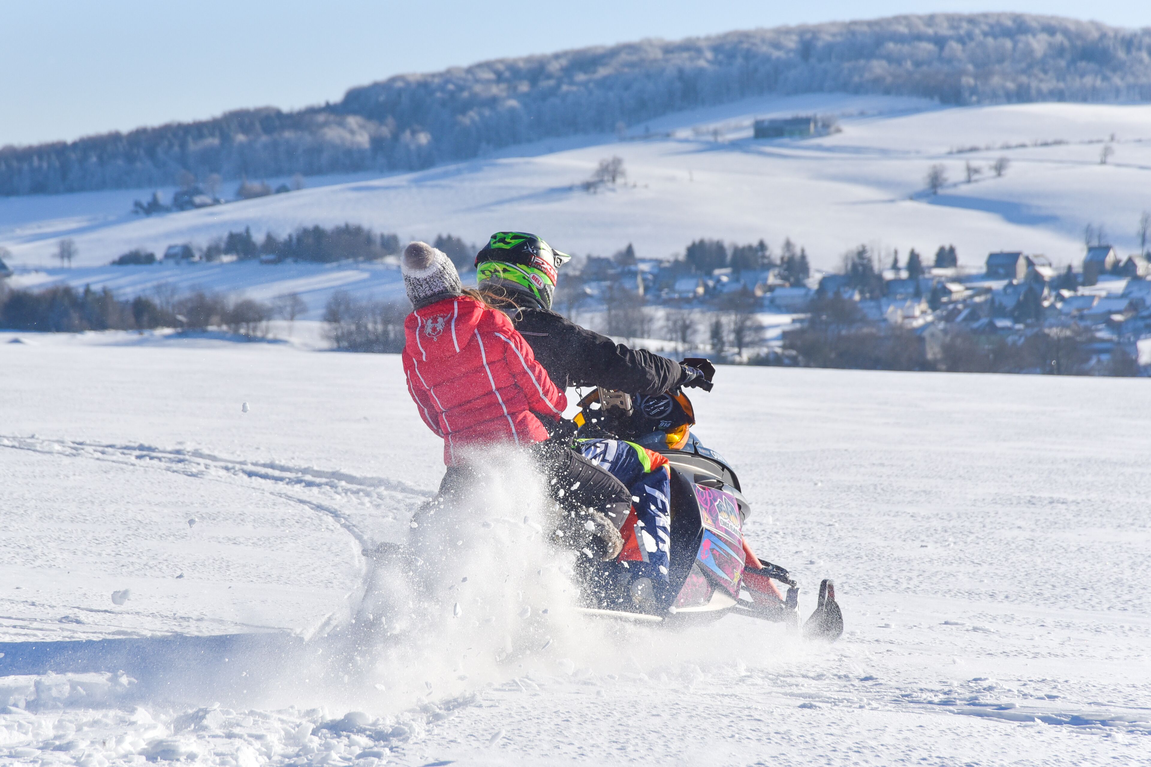 A person riding a snowmobile on a snowy landscape with houses in the background.