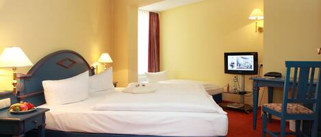 Quarto de hotel com cama, duas mesas de cabeceira com luminárias, uma televisão, uma cadeira e uma mesinha com frutas.