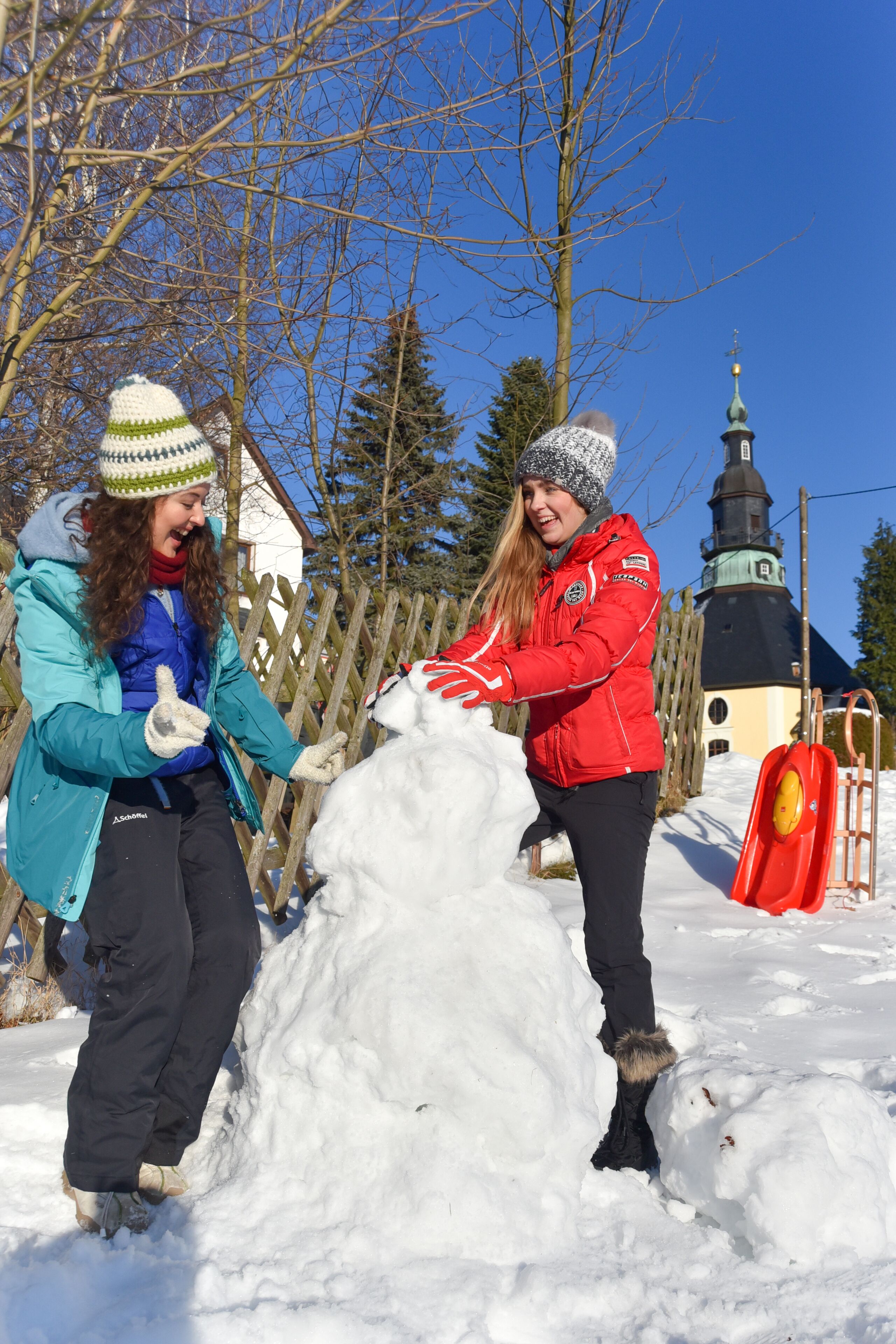Zwei Personen bauen einen Schneemann in einer verschneiten Winterlandschaft, mit einem Holzzaun und einer Kirche im Hintergrund.