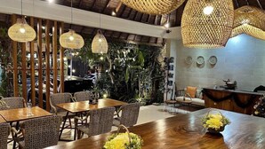 A modern dining area with wooden tables and chairs, hanging wicker lamps, and a backdrop of greenery. - Villa Puriartha (Ubud)