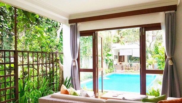 A room with a view of a swimming pool, a wicker sofa with cushions, and a wooden fence with greenery. - Villa Puriartha (Ubud)