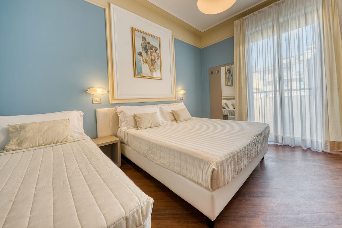 A hotel room with two beds, a large painting on the wall, and a window with sheer curtains.