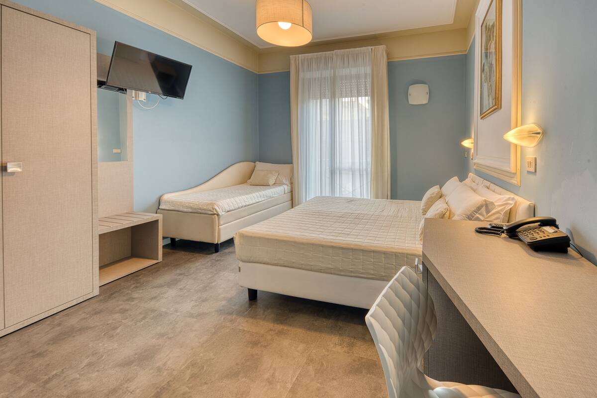 A hotel room with a bed, a desk with a telephone, a television mounted on the wall, and a wardrobe.