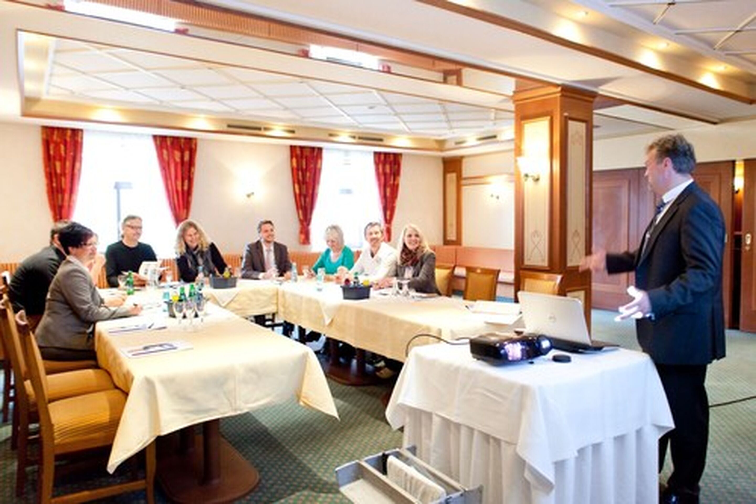 A conference room with tables set for a meeting, a speaker at the front, and a large window with curtains.