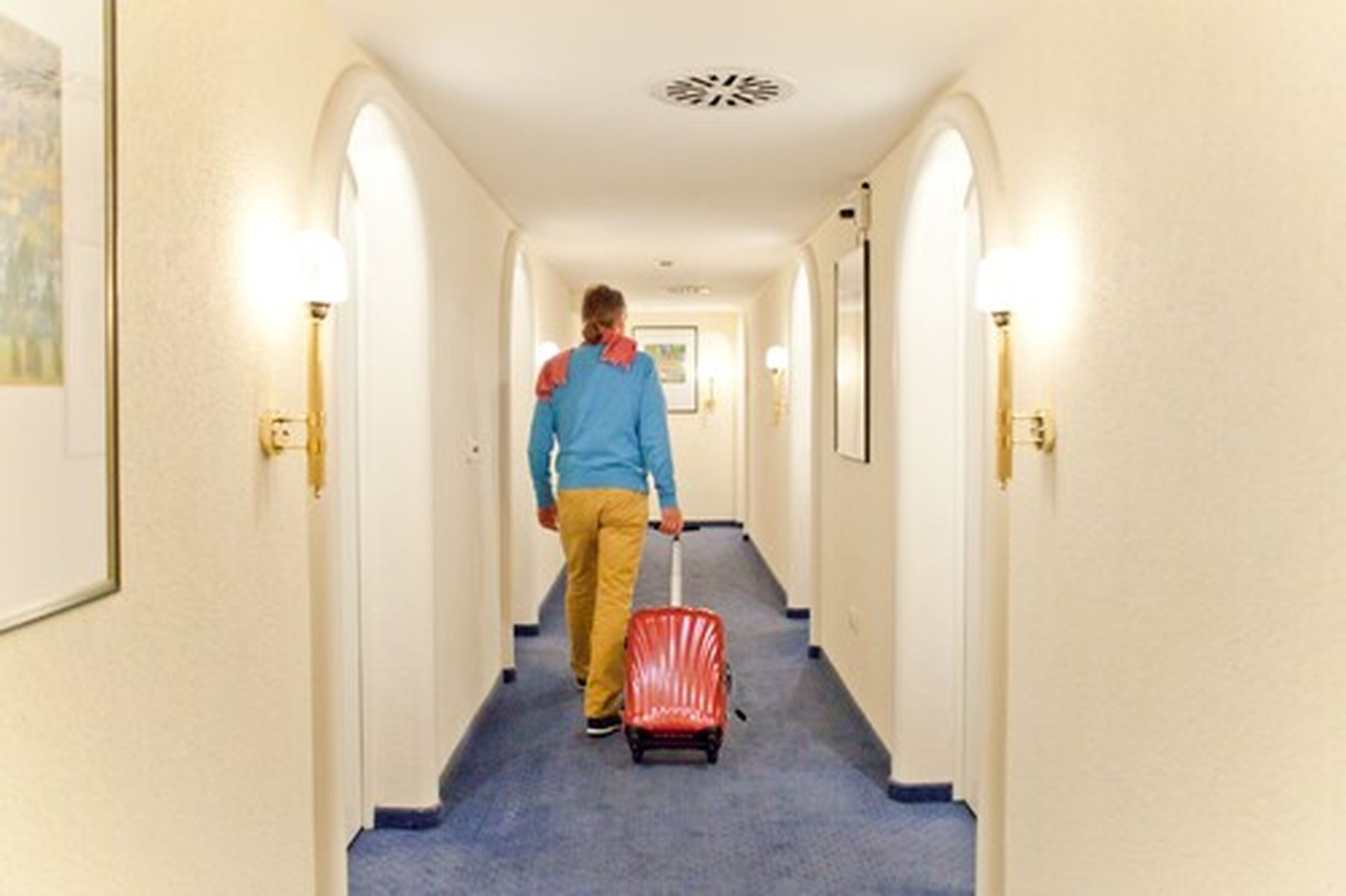 A person walking down a hotel hallway with a red suitcase.